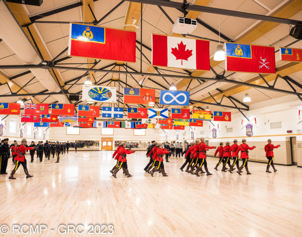 RCMP Cadets in Red Serge, marching in the hall at Depot with flags above. 
