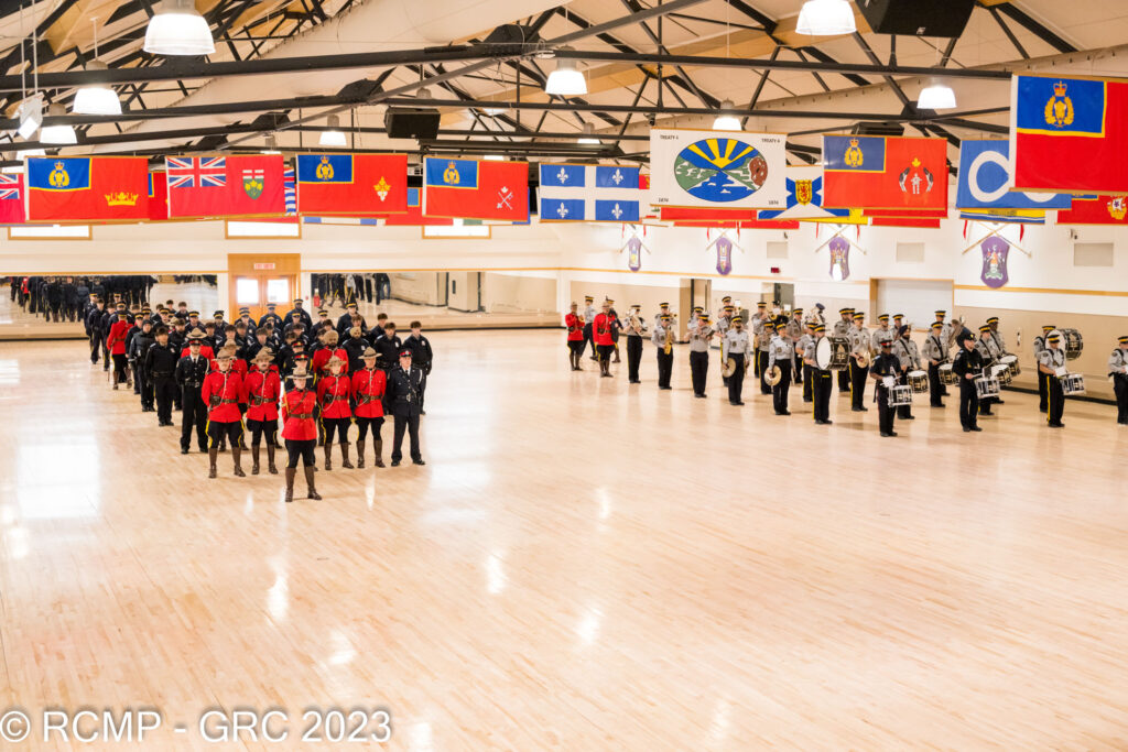 RCMP Members in training lined up in the training hall, wearing Red Serge and  the RCMP uniform. Some are holding instruments. 